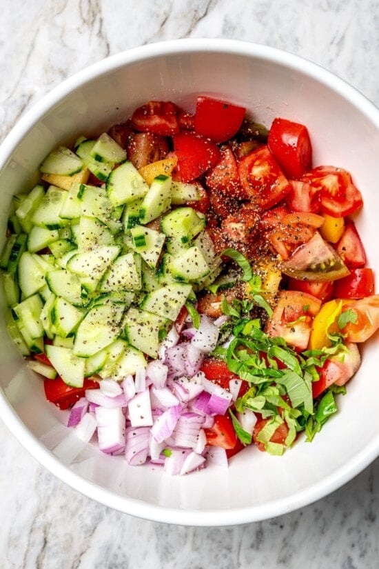 With juicy heirloom tomatoes, crisp cubes of bread, cucumber, and fresh basil, this panzanella recipe is a dish you’ll find yourself making all summer long. Serve it as a side or as a light meal. Overhead view of basil, tomatoes, red onions, and cucumbers in bowl for panzanella