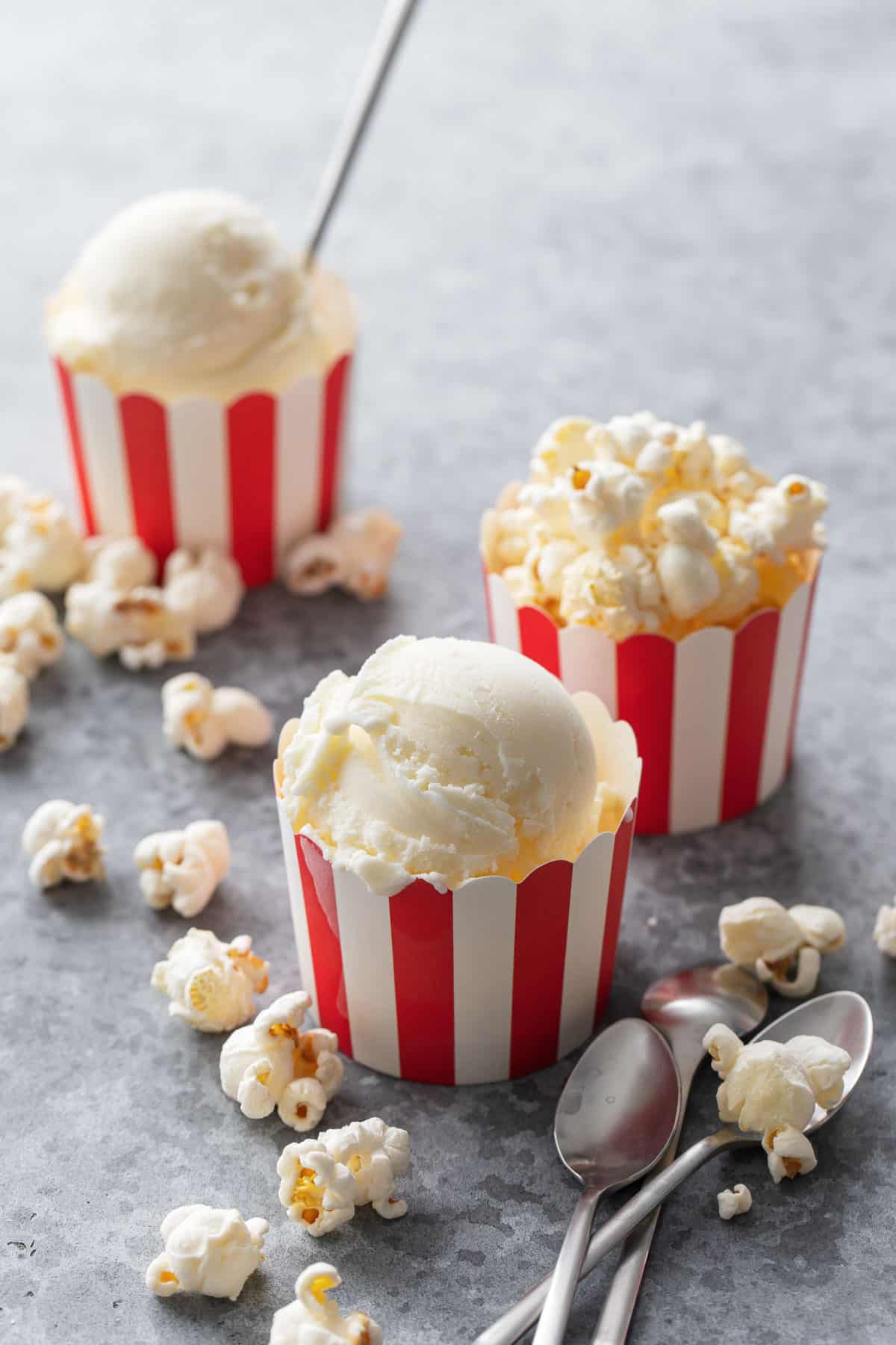 Three red and white striped containers with scoops of Buttered Popcorn Ice Cream, on a gray background with popped popcorn scattered around.