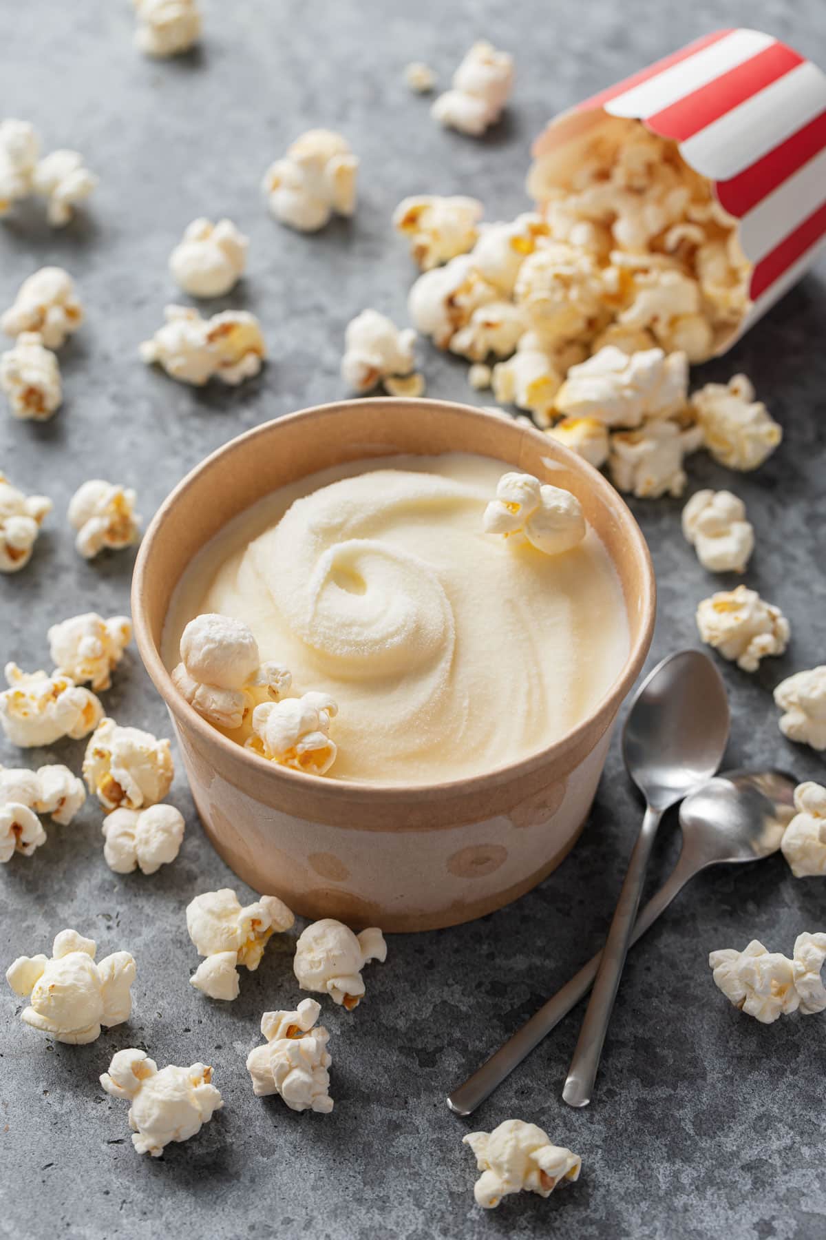 Brown paper pint container with a swirl of homemade Buttered Popcorn Ice Cream on a gray background with a spilled popcorn cup and scattered popcorn kernels.