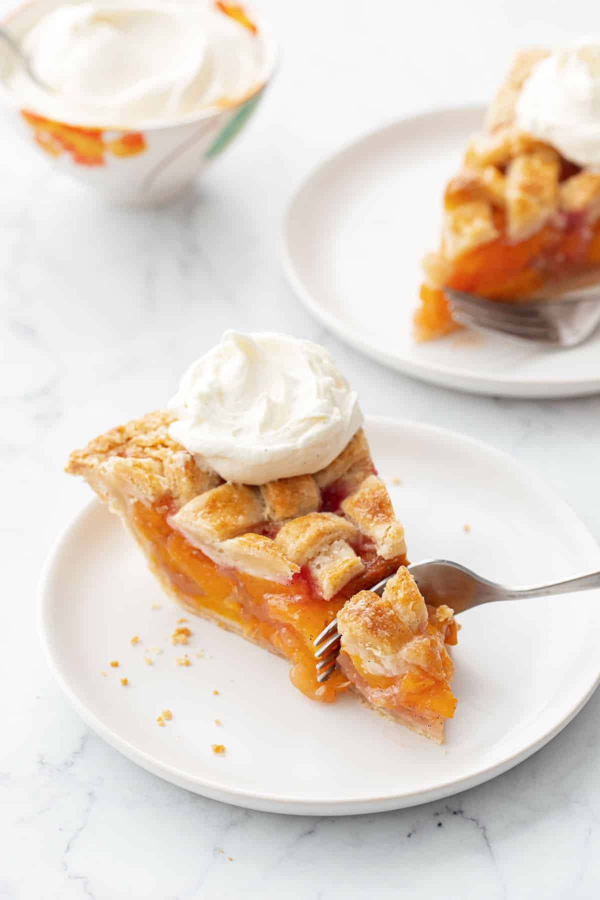 Fork cutting the tip off a slice of Vanilla Bean Peach Pie topped with a lattice crust and dollop of whipped cream.